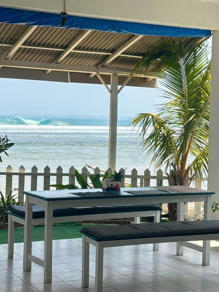 Open-air tables and benches under the awning at Lani's Resto, white picket fence and waves breaking on the Ujung Bocur reef beyond, Tanjung Setia