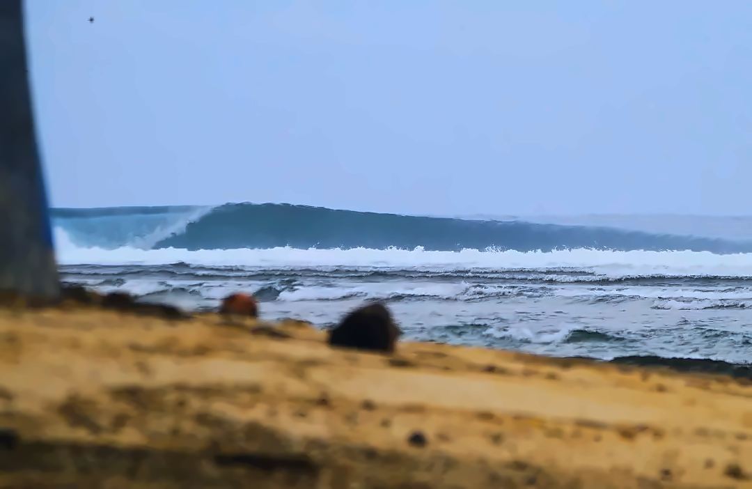 An empty wave wrapping down the Ujung Bocur reef with its lip throwing, seen from the sand at Tanjung Setia, nobody out